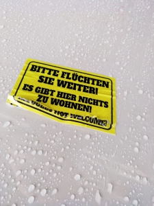 Auf gelbem Aufkleber in schwarzer Schrift steht: Bitte flüchten Sie weiter! Es gibt hier nichts zu wohnen. Refugees not welcome!