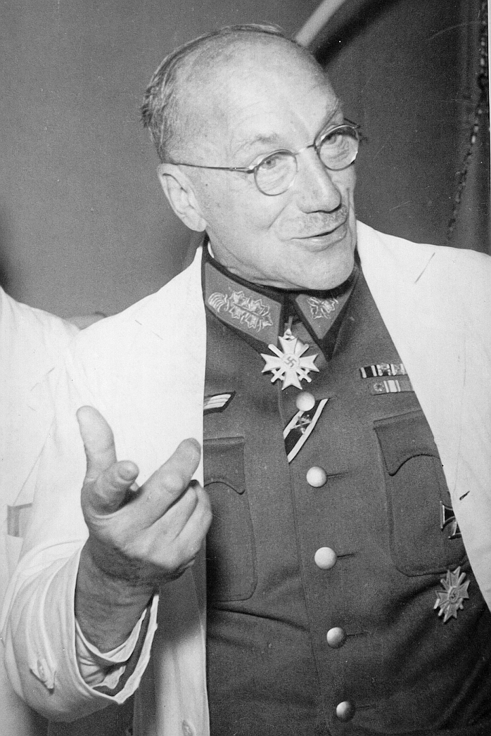 Ferdinand Sauerbruch in Wehrmachtsuniform mit diversen Orden und Abzeichen, darüber trägt er einen hellen Kittel; Mimik und Gestik lassen eine Konversation mitn einer ungezeigten Person vermuten.